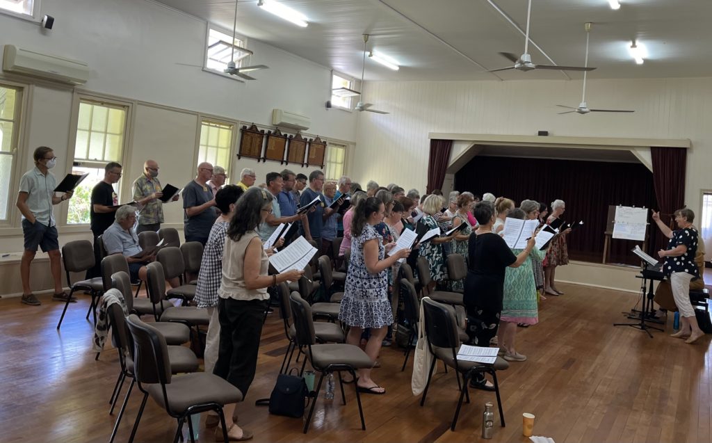 A choir in a community hall with a conductor and an accompanist. 